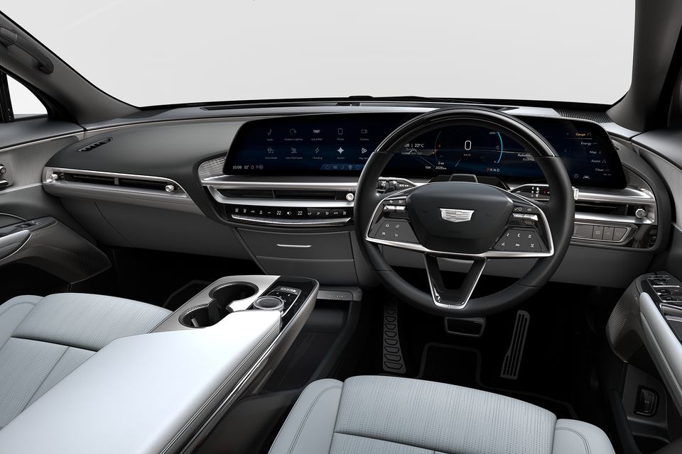 Interior of a Cadillac Lyriq showing the dashboard, large curved infotainment screen, digital instrument cluster, steering wheel, and light grey upholstery, shot from the driver's side in right-hand drive configuration