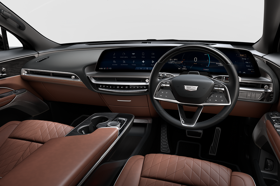 Interior of a 2027 Cadillac Lyriq showing the dashboard with a wide curved display screen, brown quilted leather seats, and Cadillac-badged steering wheel in a right-hand drive layout