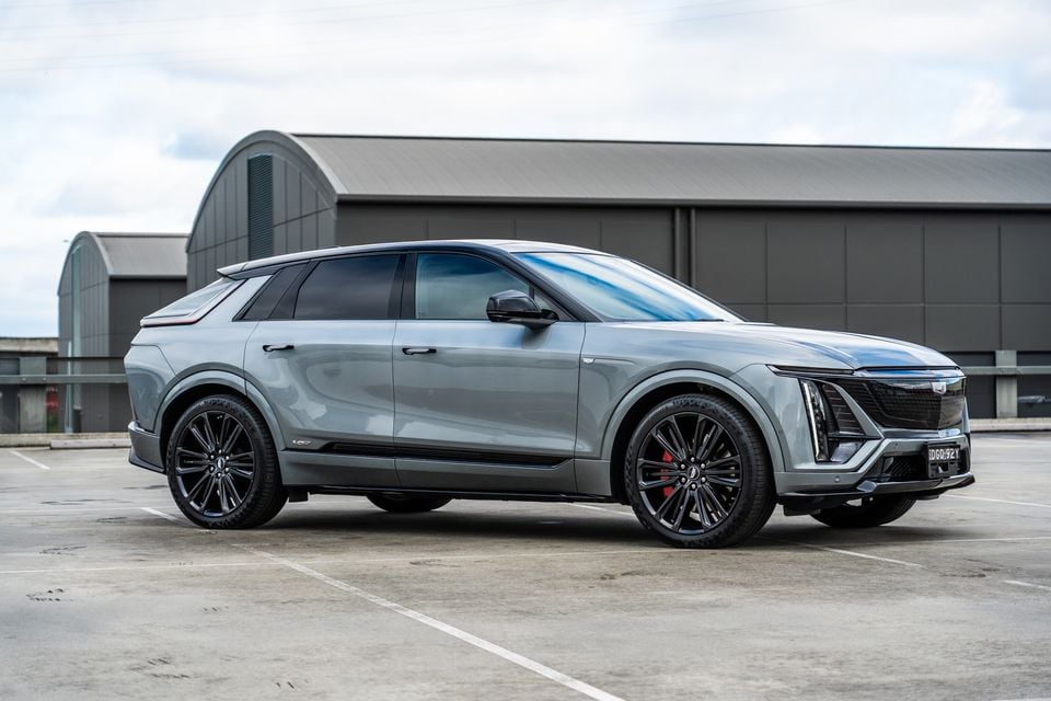 A grey Cadillac Lyriq V electric SUV parked in an open car park, seen from the front-side angle with a large industrial building in the background