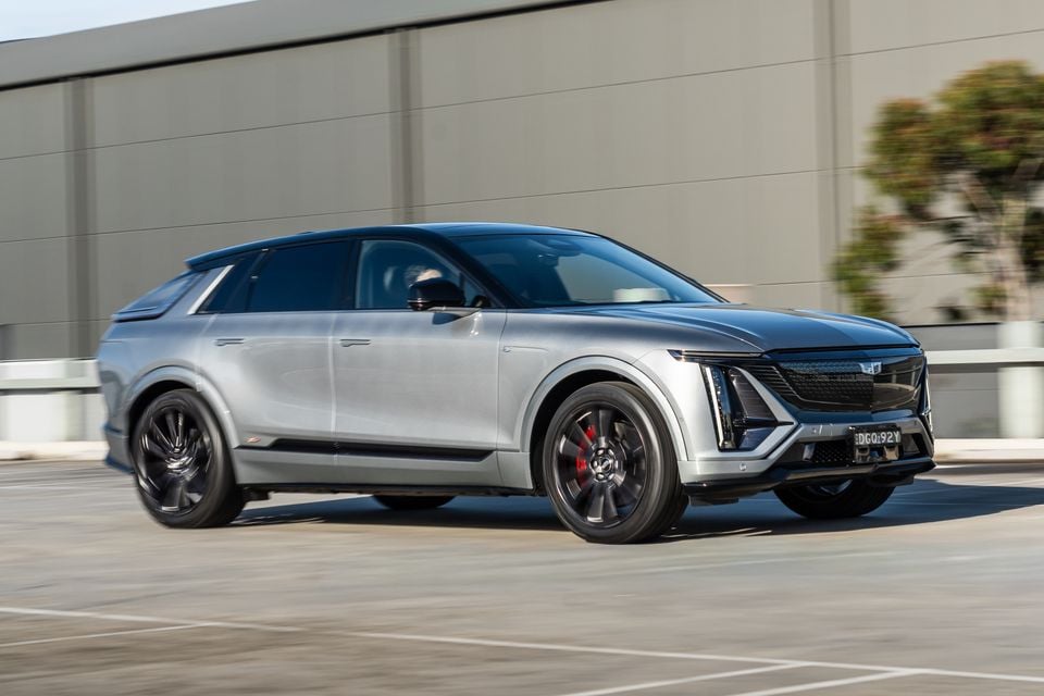 A silver 2027 Cadillac Lyriq-V electric SUV seen from the front-side, captured in motion in a car park