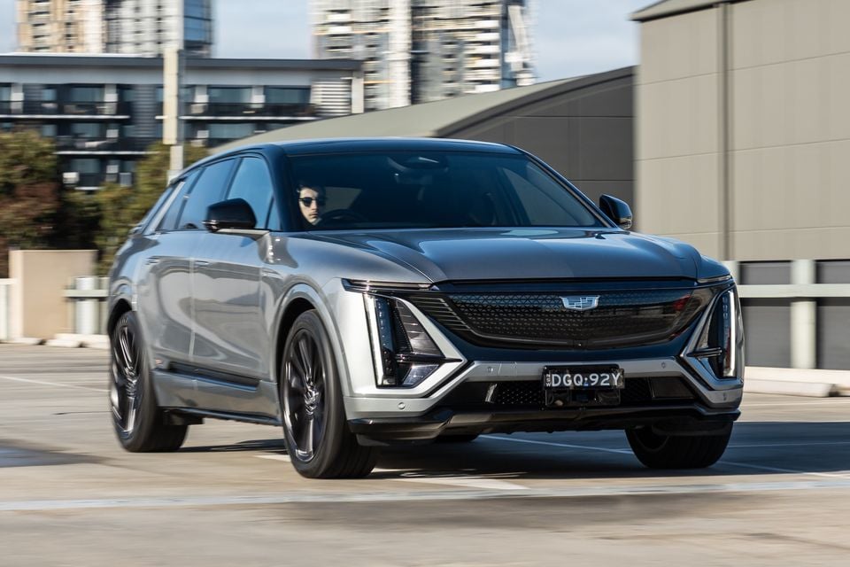A grey Cadillac Lyriq SUV seen from the front three-quarter angle driving through an urban car park area