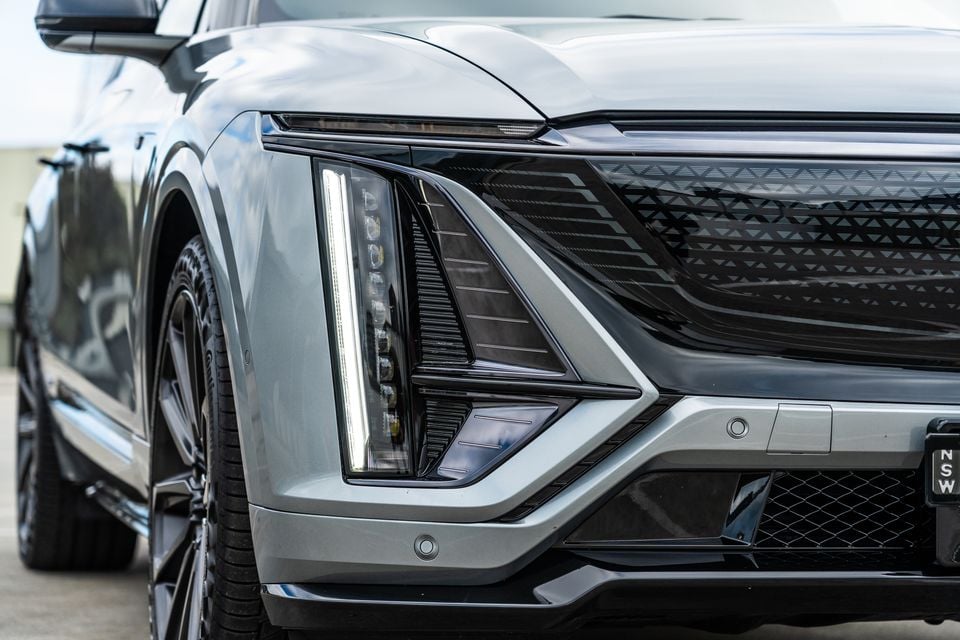 Close-up of the front corner of a grey 2027 Cadillac Lyriq, showing the angular LED headlight design and patterned grille