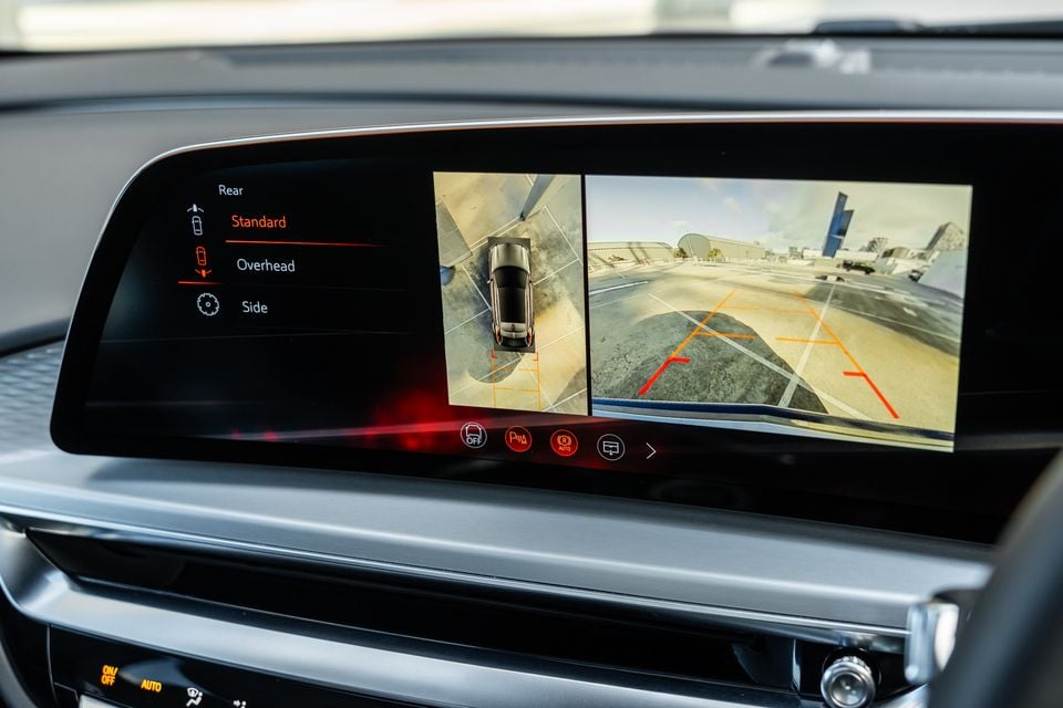 A car infotainment screen displaying a reverse camera view with an overhead bird's-eye perspective and rear camera feed, showing parking guidelines on the display.