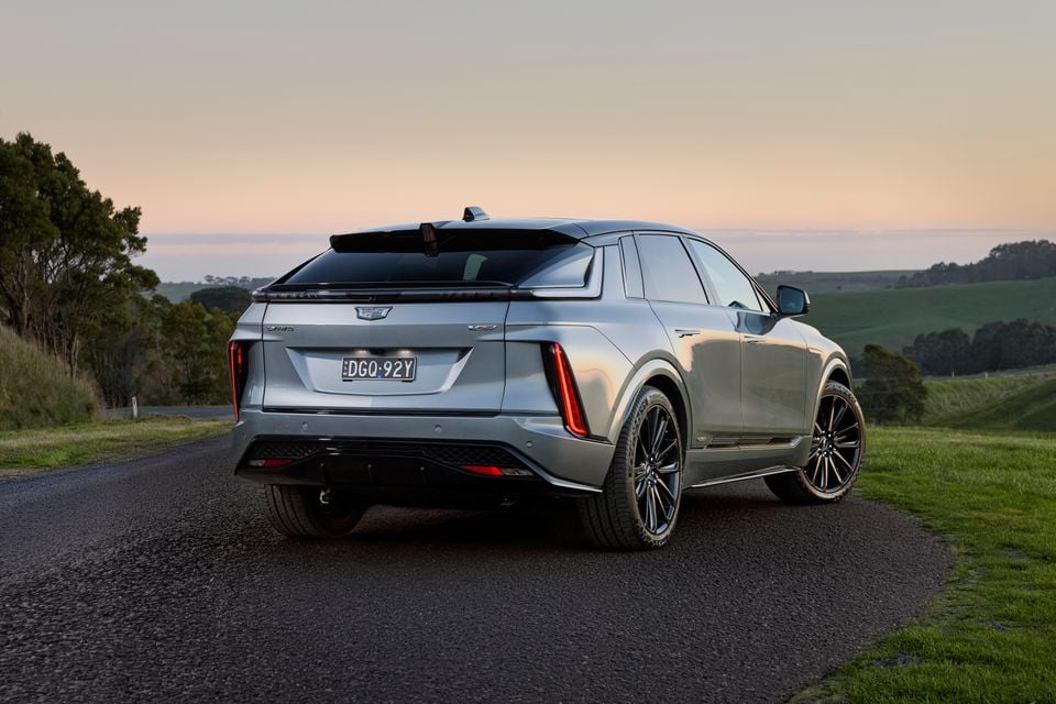 A silver 2027 Cadillac Lyriq-V SUV seen from the rear three-quarter angle on a country road at dusk