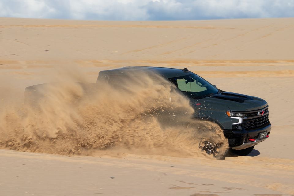 A dark Chevrolet Silverado ZR2 ute driving through sand dunes, kicking up a large spray of sand