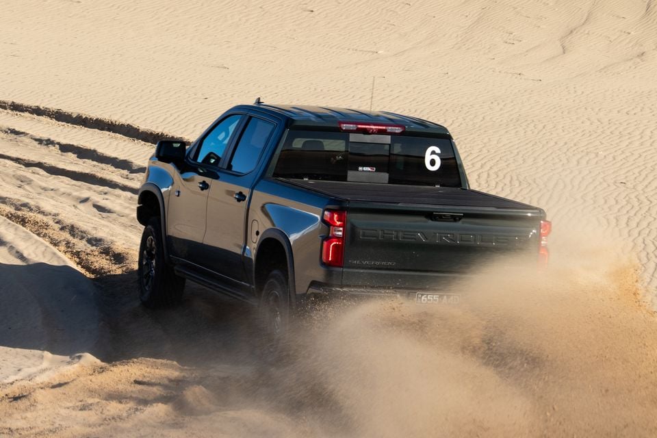 A dark grey Chevrolet Silverado ZR2 ute seen from the rear three-quarter angle, kicking up sand while driving across sand dunes