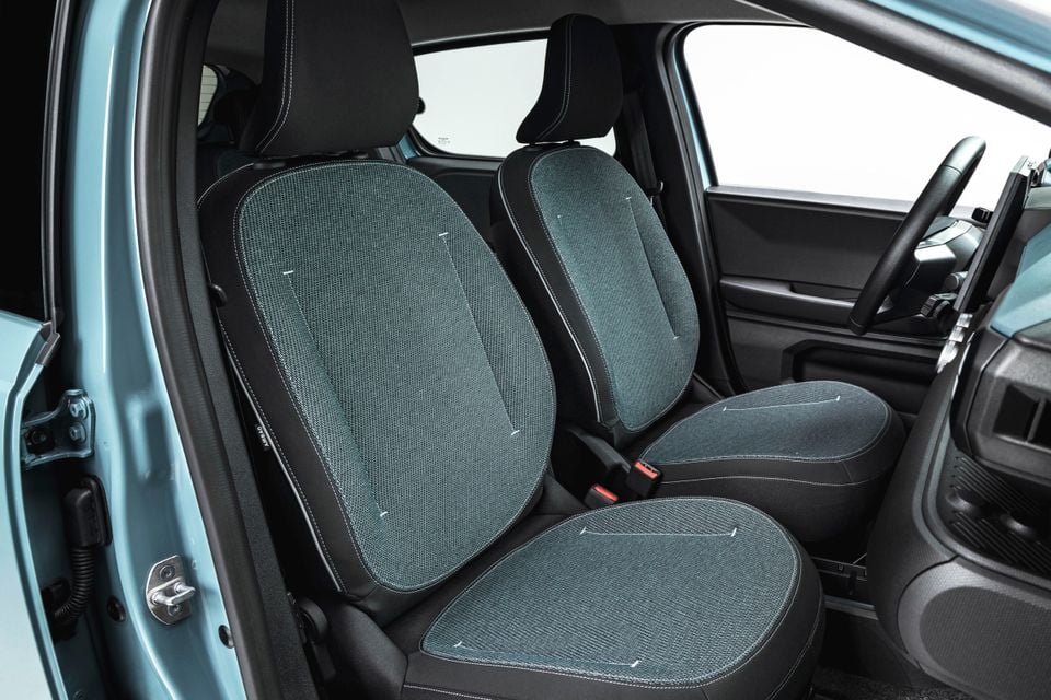 Front seats of a Dacia Spring showing teal and black fabric upholstery with white contrast stitching, viewed from the driver's door opening
