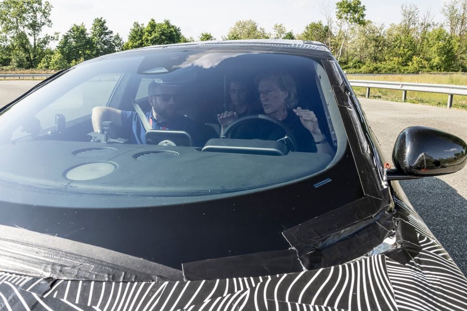 Two people sitting inside a camouflaged Ferrari prototype during a test drive on a road, viewed through the windscreen