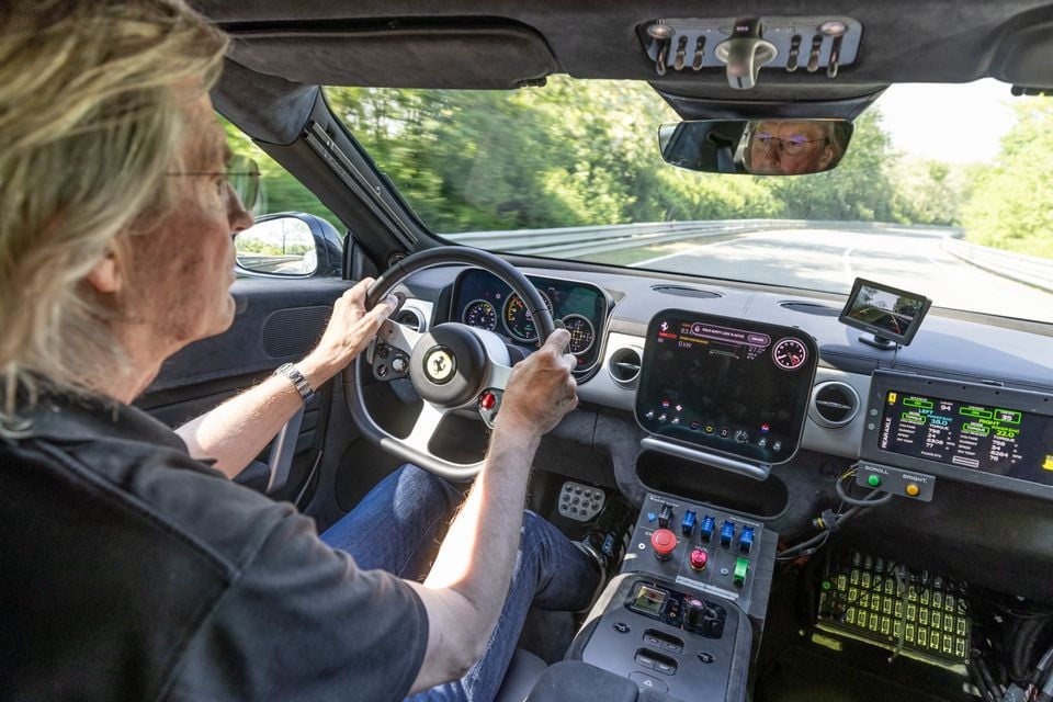 A driver at the wheel of a Ferrari prototype on a track, showing the interior with multiple screens, telemetry equipment, and an exposed console with switches and buttons