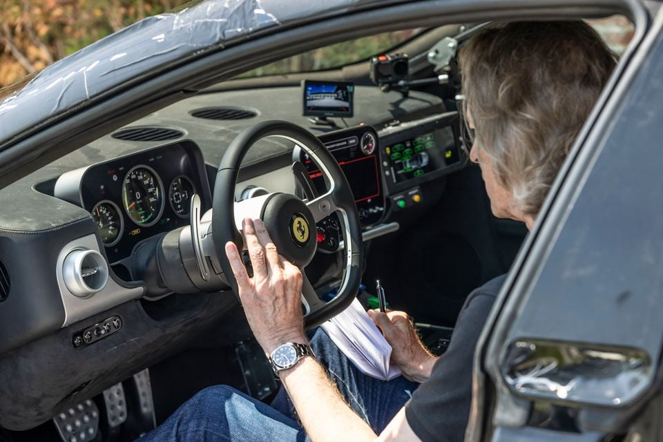 A person seated in the driver's seat of a Ferrari prototype, holding the steering wheel and taking notes, with the dashboard and instrument cluster visible