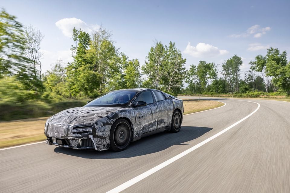 A camouflaged Ferrari Luce prototype sedan driving along a road surrounded by trees