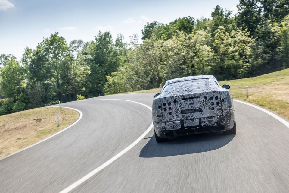 A camouflaged Ferrari Luce prototype seen from the rear while driving on a winding road surrounded by trees