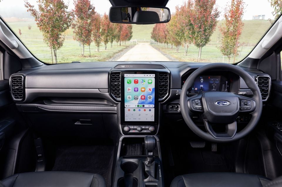 Interior dashboard view of a Ford Everest showing a large portrait touchscreen infotainment display running Apple CarPlay, steering wheel, and digital instrument cluster, with a rural tree-lined driveway visible through the windscreen