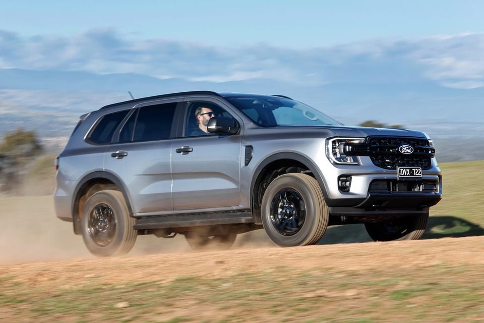 A silver Ford Everest SUV driven on a dirt track kicking up dust, seen from the front-side