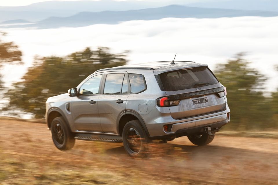 A silver Ford Everest SUV seen from the rear three-quarter angle, driving along a dirt road with a misty mountain backdrop