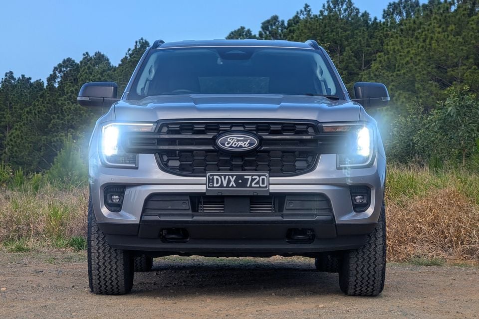 A silver Ford Everest ute seen from the front with headlights on, parked on a dirt track surrounded by bushland