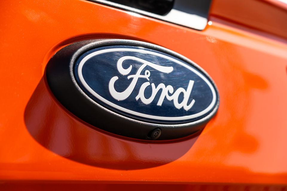 Close-up of the Ford oval badge on the tailgate of an orange Ford Ranger Wildtrak, with a reversing camera visible below