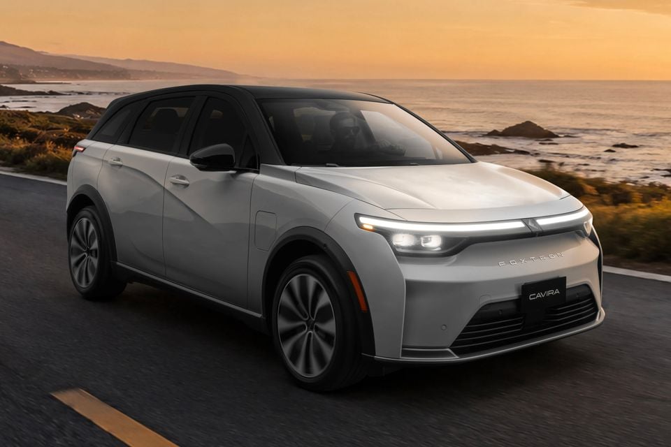 Um SUV elétrico Foxtron Cavira 2027 prateado dirigindo ao longo de uma estrada costeira ao pôr do sol, visto de frente.