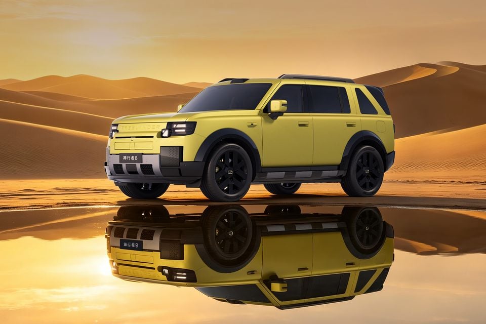A yellow Freelander 8 SUV parked on wet ground in a desert at sunset, shown from the front quarter with its reflection visible in the water.