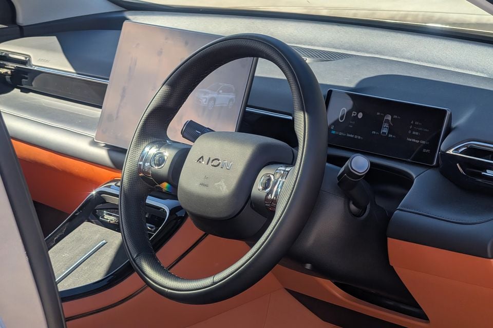 Interior of a GAC Aion V showing the steering wheel with Aion logo, large central touchscreen and digital instrument cluster, with orange and black trim