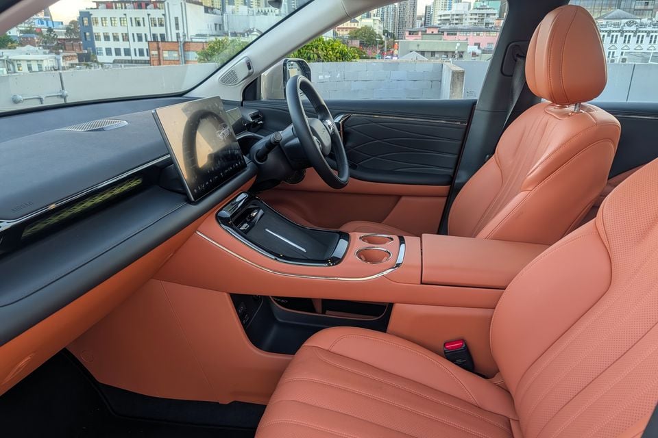 Interior of a GAC Aion V showing tan leather seats, centre console with cup holders, and a large curved infotainment touchscreen, shot from the passenger side door opening