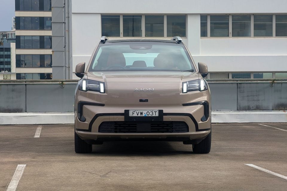 A tan GAC Aion V SUV seen from the front, parked in an open car park with city buildings in the background