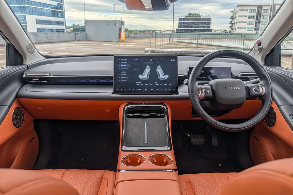 Interior of the GAC Aion V showing the orange leather cabin, large central touchscreen infotainment system, steering wheel with Aion logo, and floating centre console with cup holders
