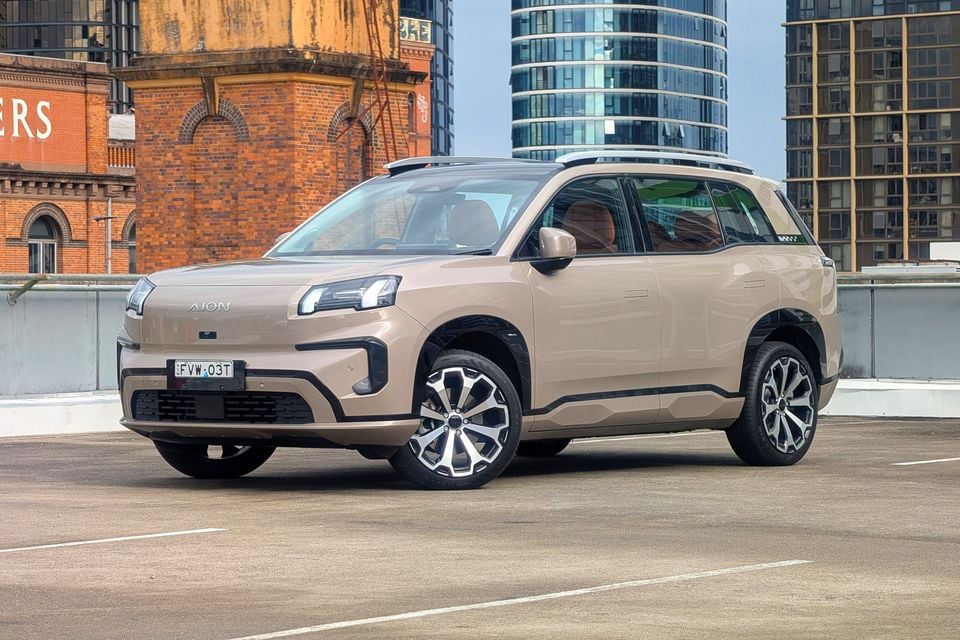 A beige GAC Aion V SUV seen from the front-side angle, parked on a rooftop car park with city buildings in the background