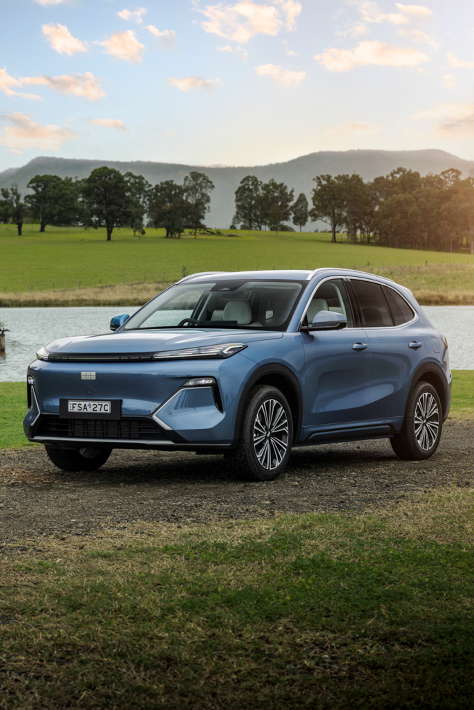 A blue 2027 Geely Starray EM-i Inspire SUV parked on gravel beside a dam, with green paddocks, trees and mountains behind
