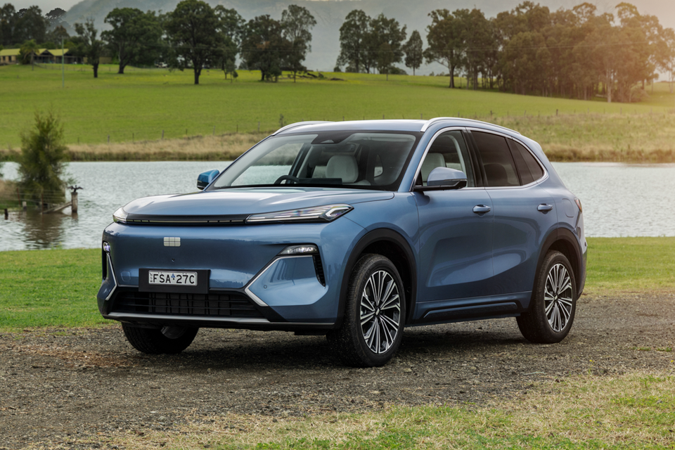 A blue 2027 Geely Starray EM-i Inspire SUV parked on gravel beside a lake, seen from the front three-quarter angle