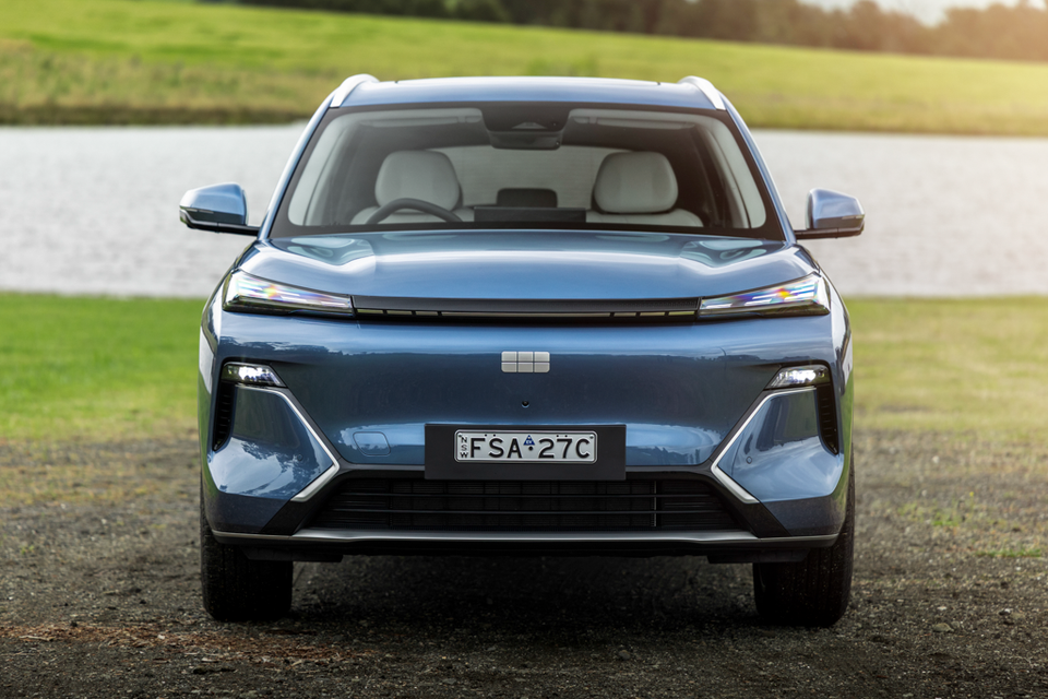 A blue 2027 Geely Starray EM-i Inspire SUV seen from the front, parked on gravel beside a lake and grassy field