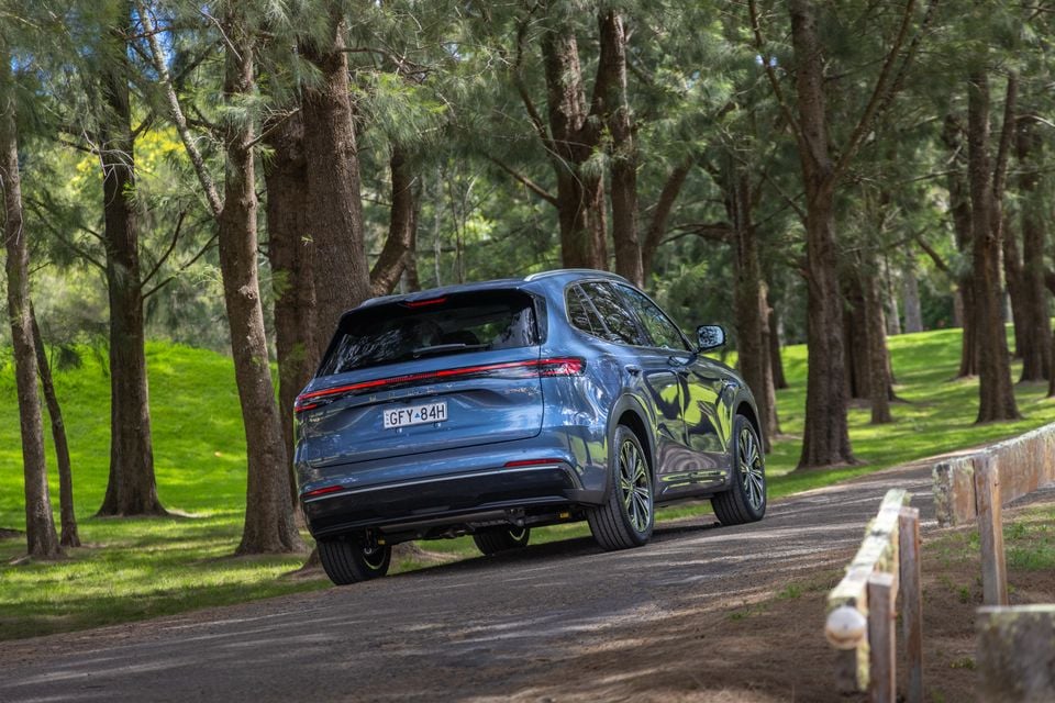 A blue 2027 Geely Starray EM-i SUV seen from the rear, driving along a gravel road through a pine forest