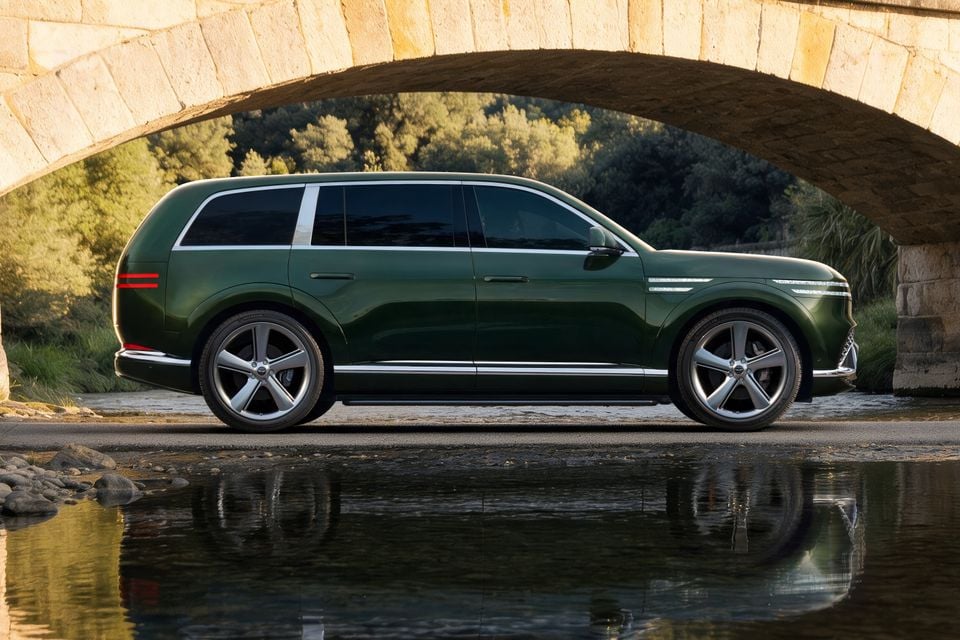 A green Genesis GV90 large SUV seen from the side, parked beneath a stone arch bridge near a reflective body of water