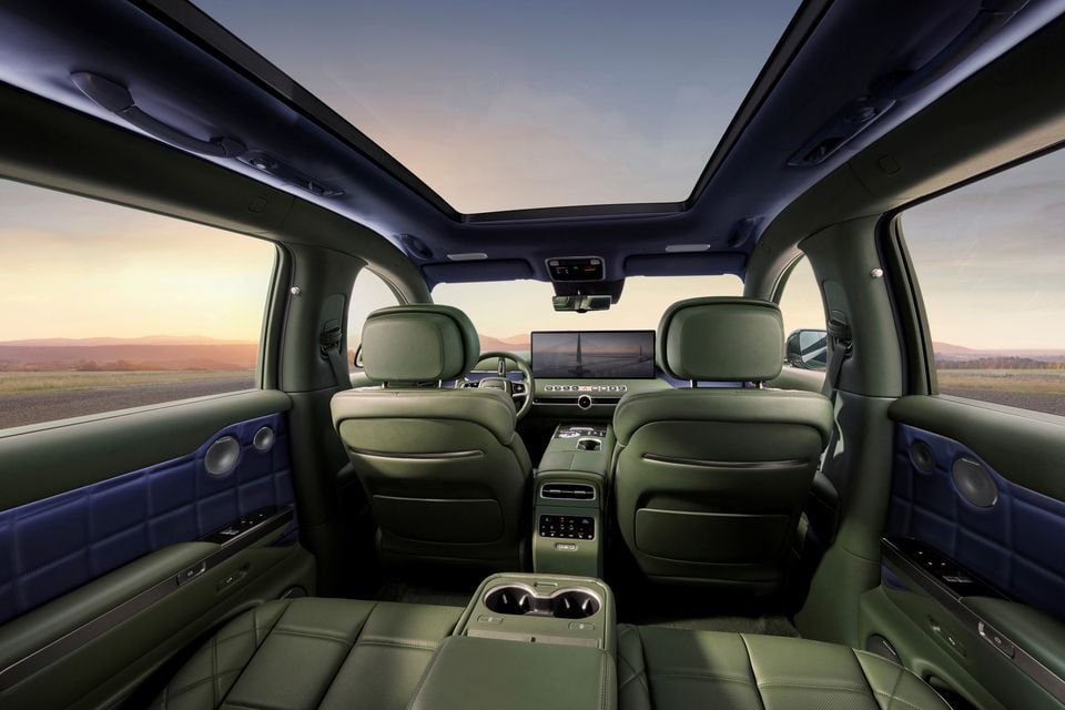 Interior of the 2027 Genesis GV90 viewed from the rear, showing green leather seats, a panoramic sunroof, large central infotainment screen, and two-tone blue and green cabin trim