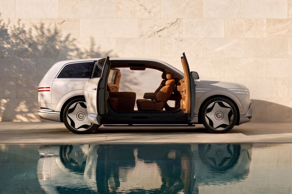 A silver 2027 Genesis GV90 Neolun luxury SUV seen from the side with coach doors open, revealing a tan leather interior, reflected in a pool of water beside a stone wall.