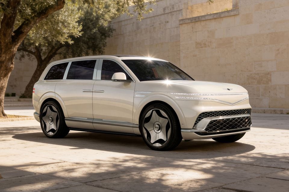 A cream/white 2027 Genesis GV90 Neolun large SUV seen from the front-side angle, parked on a stone courtyard beside a sandstone wall