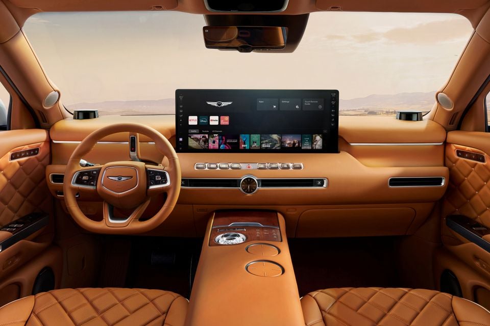 Interior of a Genesis GV90 showing tan quilted leather seats, a wide touchscreen infotainment display with streaming apps, analogue clock, and centre console
