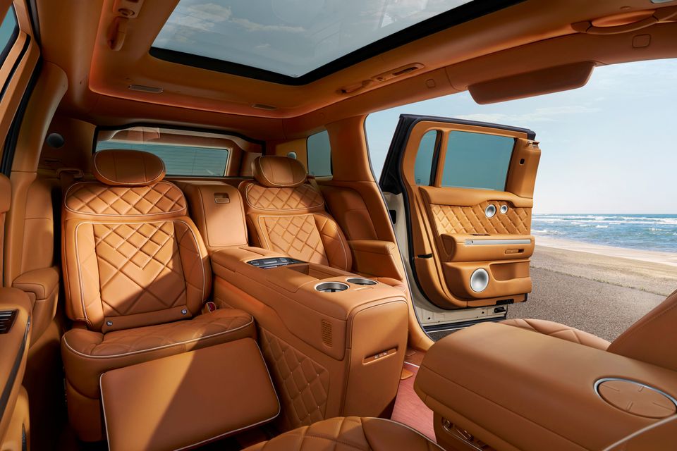 Luxurious tan leather rear interior of the 2027 Genesis GV90 Neolun, showing diamond-quilted reclining seats, centre console with cup holders, panoramic sunroof, and coach door open to a beachside backdrop.