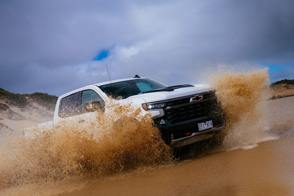 A white Chevrolet Silverado ZR2 ute ploughing through a muddy water crossing on a beach, sending a large spray of muddy water to the sides