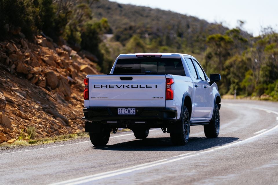 A white Chevrolet Silverado ZR2 ute seen from the rear, driving along a winding road with rocky terrain and bushland in the background