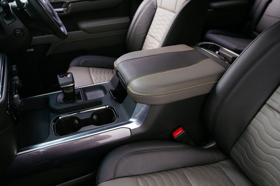 Interior view of a car's centre console showing the leather-trimmed armrest with yellow contrast stitching, cup holders, gear selector, and quilted front seats
