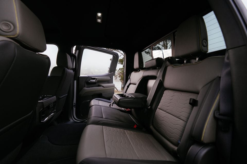 Rear interior cabin of a 2027 Chevrolet Silverado ZR2 showing black leather rear seats with yellow contrast stitching and a fold-down centre armrest, with the rear door open