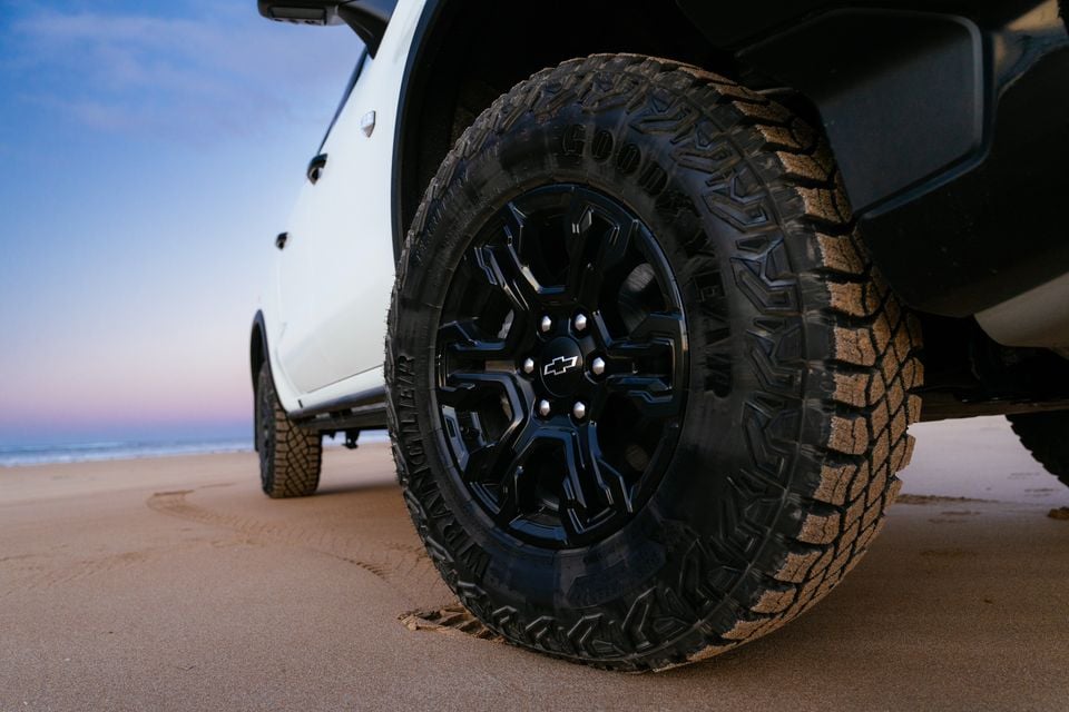 Close-up of a Goodyear Wrangler all-terrain tyre on a black alloy wheel with a Chevrolet badge, fitted to a white Chevrolet Silverado ZR2 parked on a beach at dusk