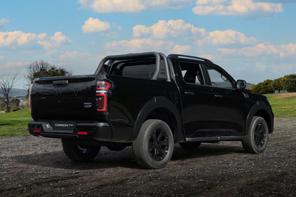 A black GWM Cannon PHEV dual-cab ute seen from the rear three-quarter angle on a gravel surface with a rural backdrop