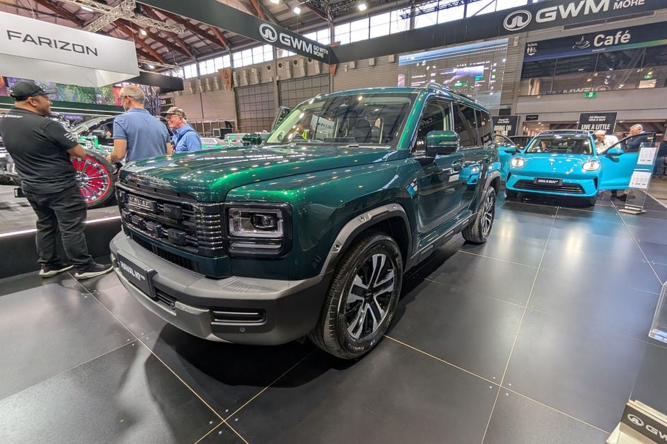 A dark green GWM Haval H7 SUV displayed at a motor show, seen from the front-side angle, with a blue ORA 5 SUV visible in the background at the GWM stand.