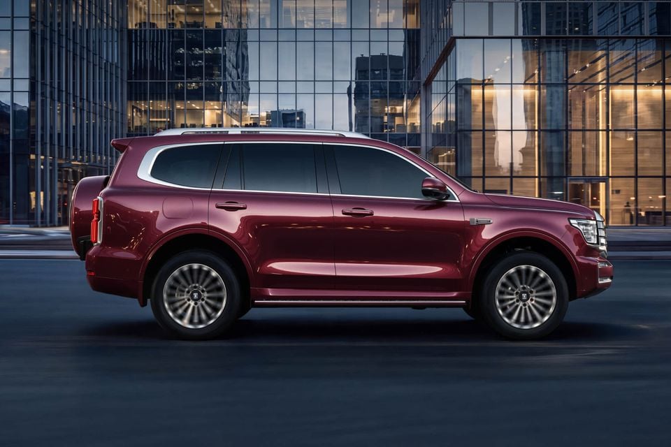A red 2027 GWM Tank 500 SUV seen side-on, driving past glass office buildings at dusk