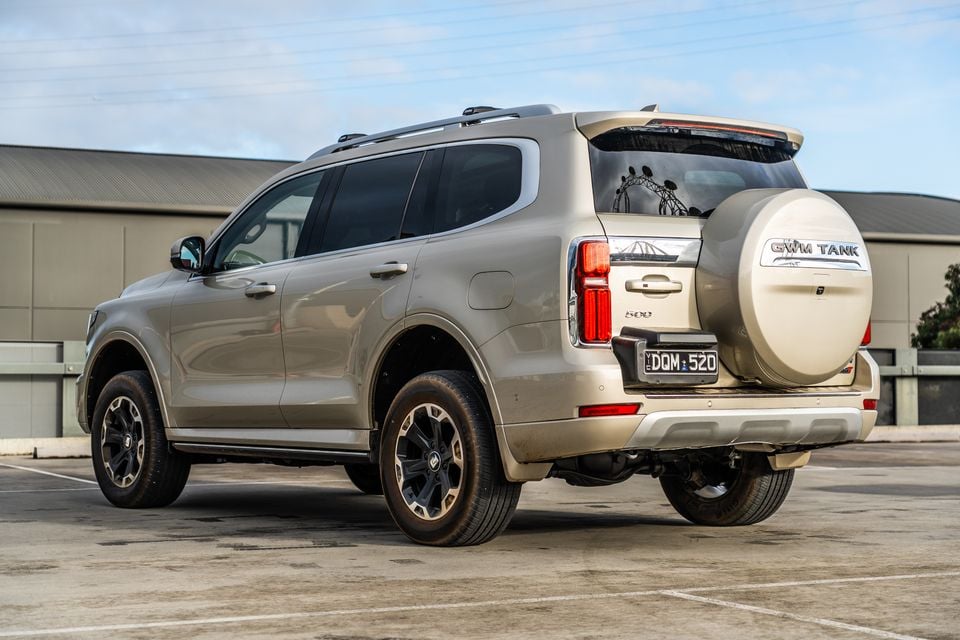 A GWM Tank 500 large SUV seen from the rear three-quarter angle in a car park, showing the exterior spare wheel and gold/champagne paintwork