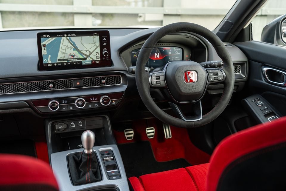 Interior of a Honda Civic Type R showing the flat-bottom suede steering wheel with red Honda badge, touchscreen navigation display, manual gear shifter, and red sport seats