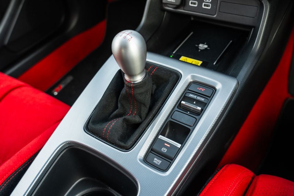 Close-up of the centre console in a Honda Civic Type R, showing a short-throw manual gear shifter with red stitching, drive mode selector including a +R button, electronic parking brake, and brake hold button, with red Alcantara seats visible on either side.