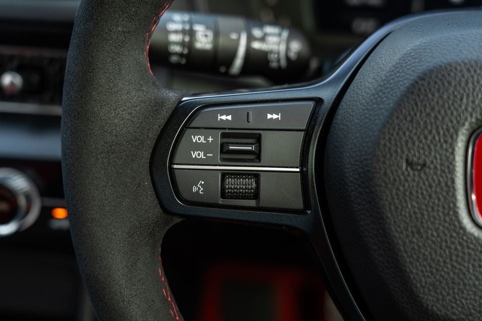 Close-up of the steering wheel controls on a Honda Civic Type R, showing media playback, volume, and voice command buttons on an Alcantara-wrapped wheel with red stitching.